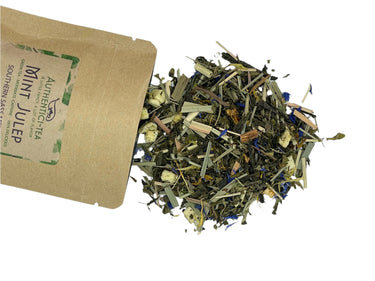 A brown paper packet labeled “Mint Julep Artisan Tea – Southern Charm in a Teacup 🌿🍋🥃” spills loose dried tea leaves, blue petals, spearmint, peppermint, and bourbon-flavored herbs onto a white surface.